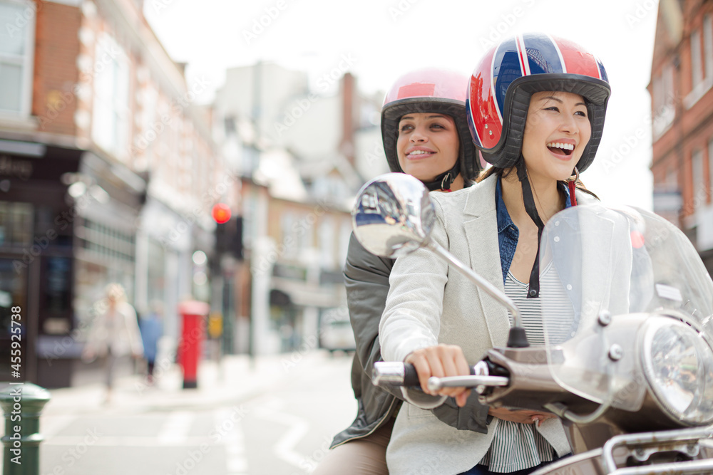 Smiling young women friends wearing helmets, riding motor scooter on urban street Smiling young women friends wearing helmets, riding motor scooter on urban street