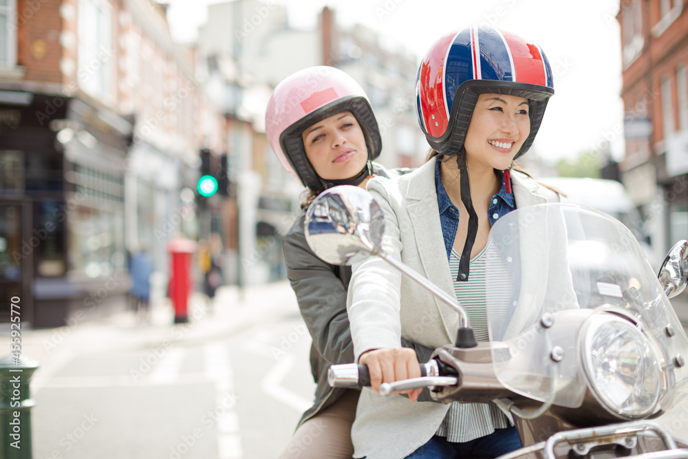 Smiling young women friends wearing helmets, riding motor scooter on urban street Smiling young women friends wearing helmets, riding motor scooter on urban street