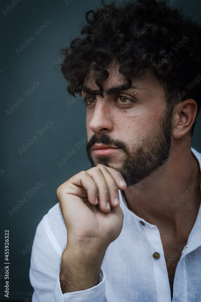 portrait of a young man with beard and curly hair, holding his chin with his hand and looking thoughtful portrait of a young man with beard and curly hair, holding his chin with his hand and looking thoughtful