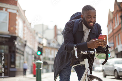 Young businessman commuting with bicycle, texting with cell phone on sunny urban street