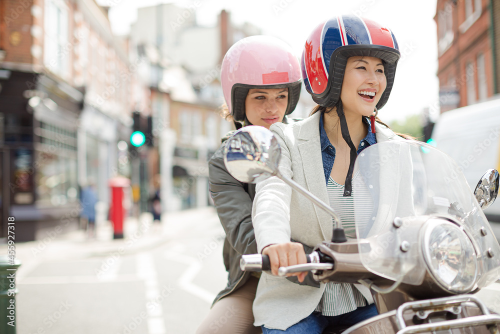 Smiling young women friends wearing helmets, riding motor scooter on urban street Smiling young women friends wearing helmets, riding motor scooter on urban street