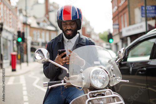Smiling young businessman in helmet on motor scooter texting with cell phone on urban street