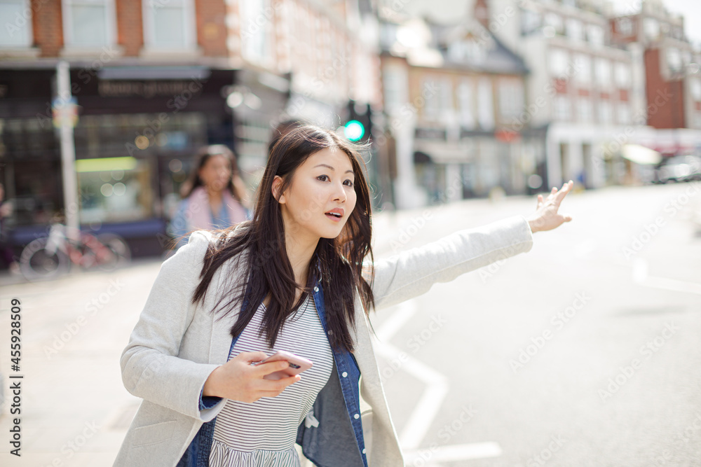 Fototapeta premium Young businesswoman with cell phone hailing taxi on sunny urban street