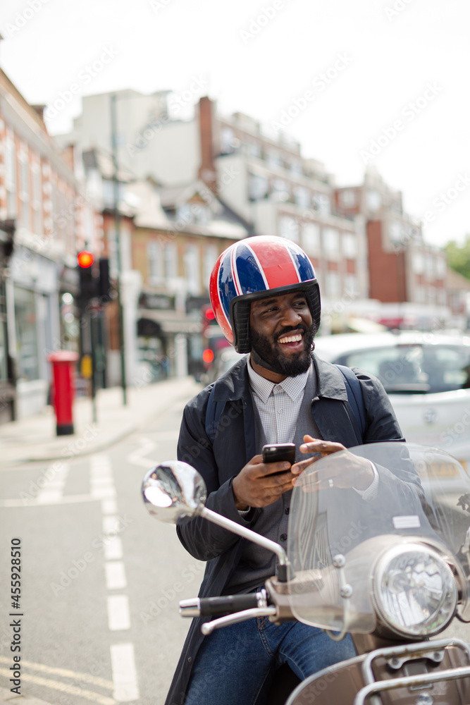 Smiling young businessman in helmet on motor scooter texting with cell phone on urban street Smiling young businessman in helmet on motor scooter texting with cell phone on urban street