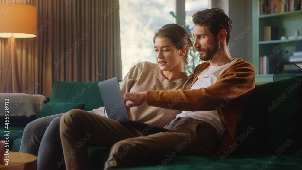 Young Couple Use Laptop Computer, while Sitting on a Couch in the Cozy Stylish Apartment. Boyfriend and Girlfriend Shopping on Internet, Watching Videos and Streaming Service. Young Couple Use Laptop Computer, while Sitting on a Couch in the Cozy Stylish Apartment. Boyfriend and Girlfriend Shopping on Internet, Watching Videos and Streaming Service.