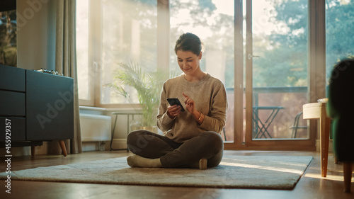 Smiling Young Woman Using Smartphone at Home, does Remote Work. Beautiful Girl Sitting on the Floor Uses Mobile Phone Internet, e-Shopping, Order Products Online, Post on Social Media. Sunny Home