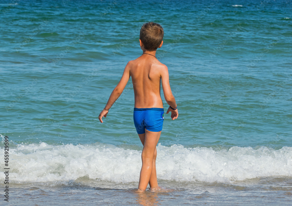 A little boy sits on the edge of the sea and admires the waves. Children on summer vacation. Rear view. A little boy sits on the edge of the sea and admires the waves. Children on summer vacation. Rear view.