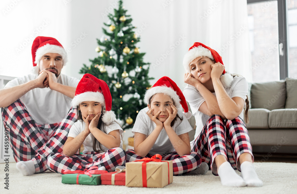 Fototapeta premium family, winter holidays and people concept - unhappy mother, father and two daughters in santa hats opening christmas gifts at home