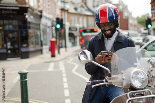 Smiling young businessman in helmet on motor scooter texting with cell phone on urban street