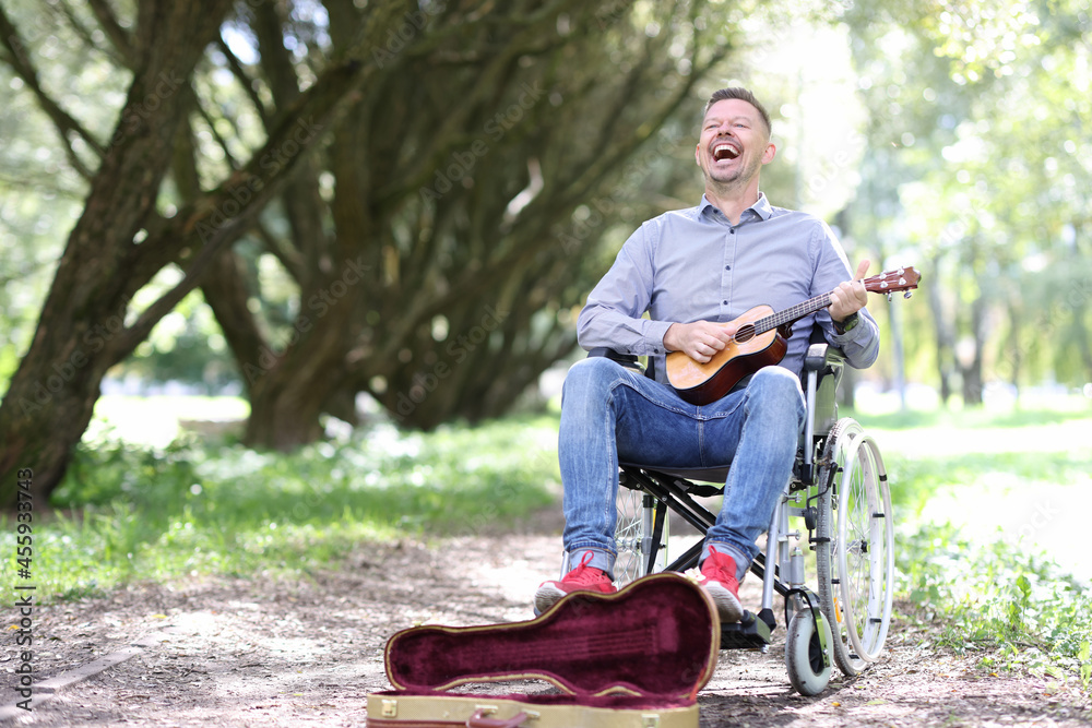 Fototapeta premium Disabled man in wheelchair plays guitar in park and makes money