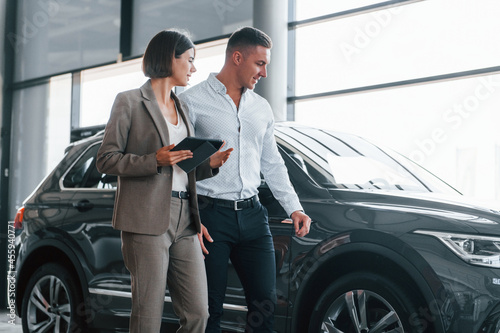 Woman in formal wear helping customer with choice of the automobile