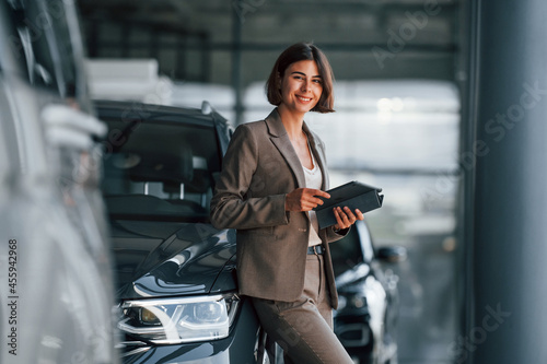 With tablet in hands. Woman is indoors near brand new automobile indoors