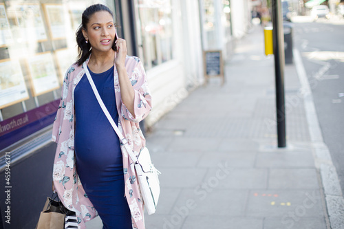 Pregnant woman talking on cell phone, walking along urban storefront