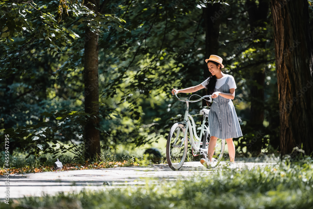 Obraz premium happy asian woman in striped sundress walking with bicycle in park