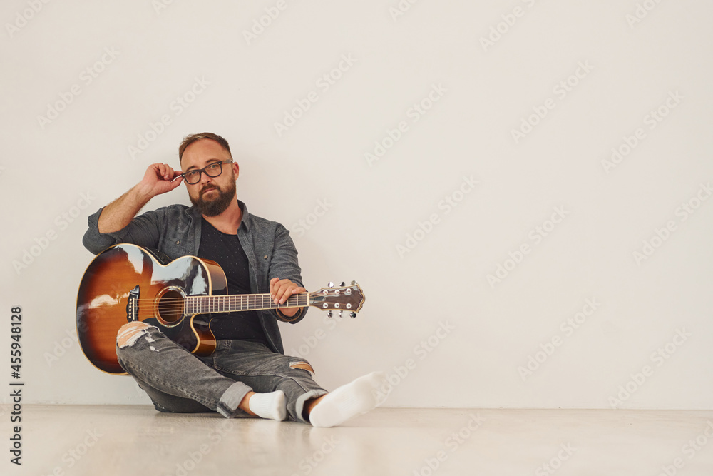 Sitting against wall. Man in casual clothes and with acoustic guitar is indoors
