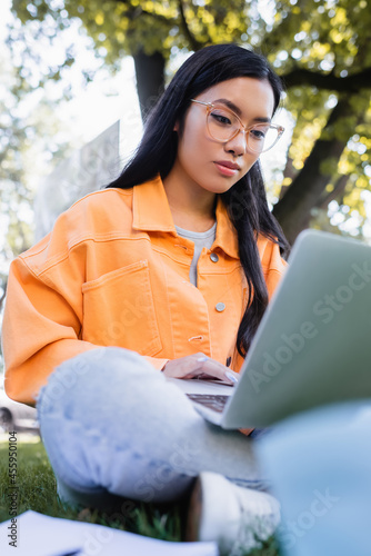 asian student in eyeglasses using laptop in park, blurred foreground