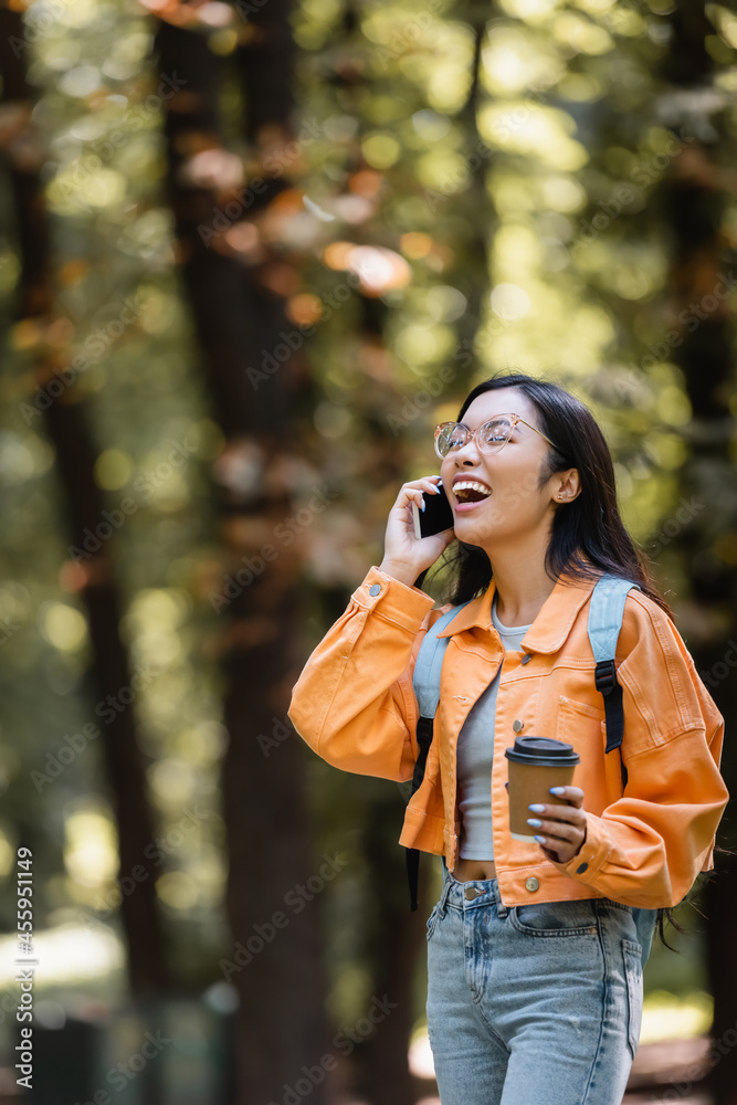 asian student laughing with closed eyes while talking on smartphone outdoors
