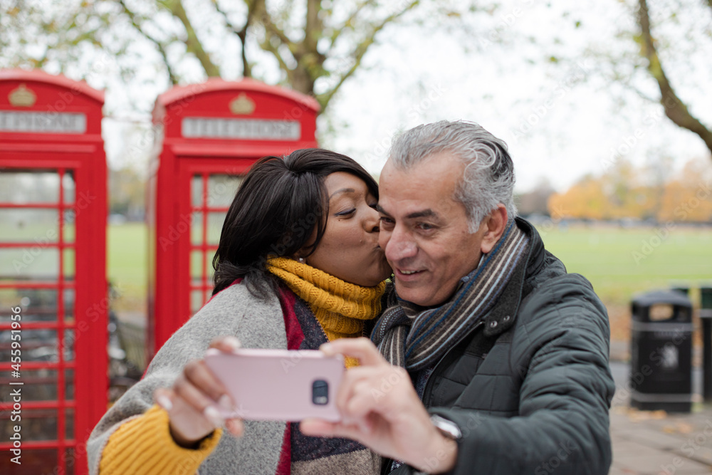 Smiling senior couple taking selfie in autumn park in front of red telephone booths Smiling senior couple taking selfie in autumn park in front of red telephone booths