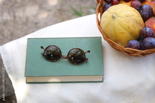 Vintage basket full of various fruit, hardback book and retro sunglasses on picnic basket. Selective focus.