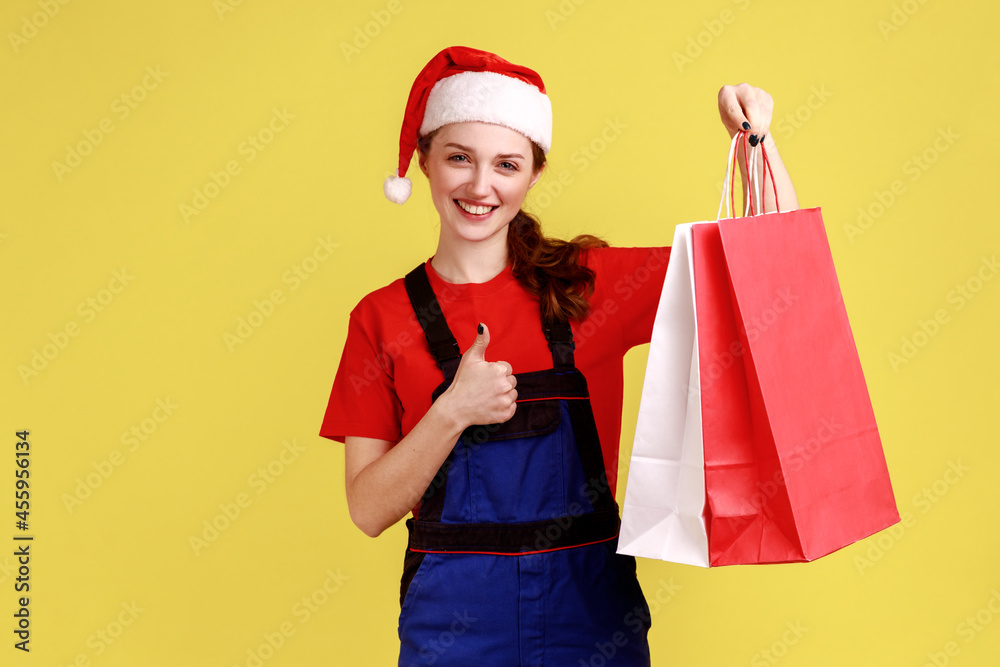 Happy female courier holding shopping bags and showing thumb up, recommend delivery service, wearing blue overalls and santa claus hat. Indoor studio shot isolated on yellow background. Happy female courier holding shopping bags and showing thumb up, recommend delivery service, wearing blue overalls and santa claus hat. Indoor studio shot isolated on yellow background.