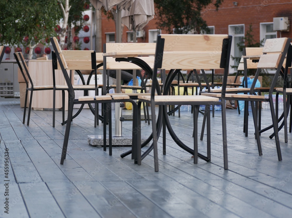 An open-air cafe is preparing to receive guests. New furniture for the cafe is installed. Tables and chairs made of black metal and light wood on a dark gray floor.
