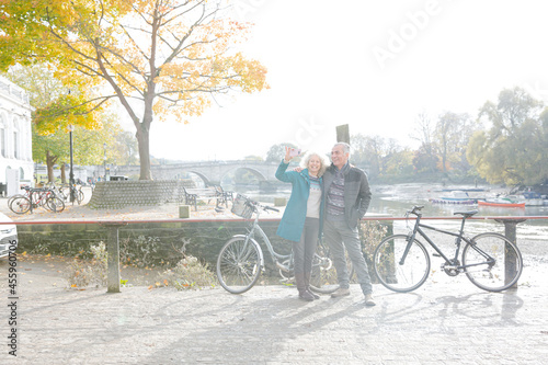 Fototapeta Naklejka Na Ścianę i Meble -  Senior couple with bicycles taking selfie at autumn river