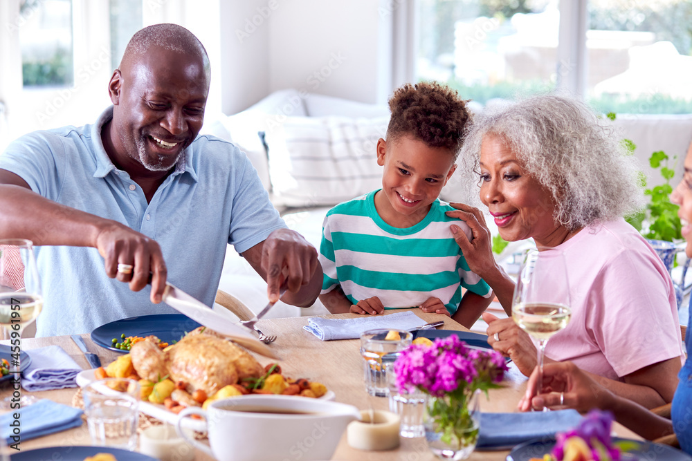 Grandfather Carving As Multi Generation Family Sit Around Table At Home And Enjoy Eating Meal Grandfather Carving As Multi Generation Family Sit Around Table At Home And Enjoy Eating Meal