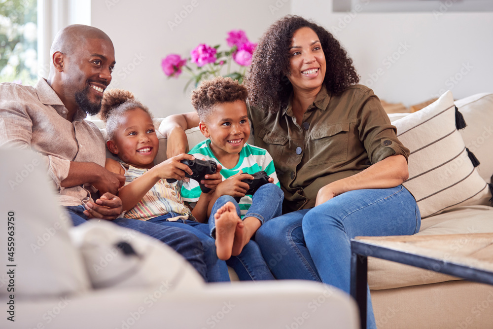 Parents Sitting On Sofa With Children At Home Playing Video Game Together