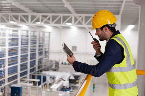 Male supervisor with digital tablet talking, using walkie-talkie on platform in factory