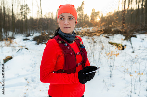 Female Runner Holding Mobile Looking for Way Out of Woods in Winter. Sportswoman Looking Around Forest Checking Location on Cellphone On Cold Winter Day.