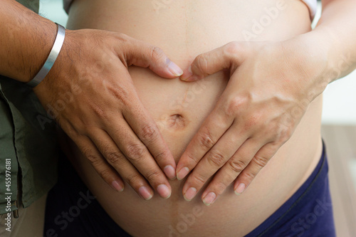 Man holding belly of his pregnant wife making heart. Man and wife making heart with their hands, closeup. Heart of hands by multiethnic couple on pregnant belly.