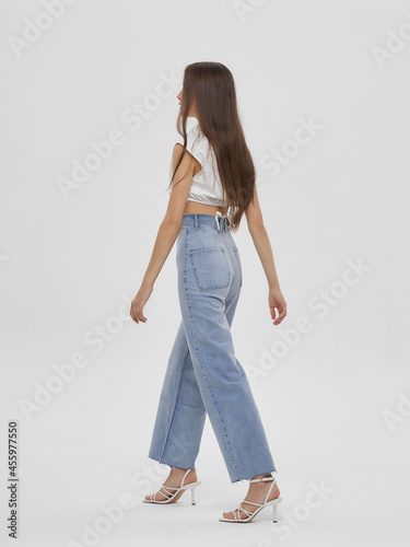 Young elegant woman with long straight hair and natural make-up in white blouse and blue jeans standing and posing on bright grey background