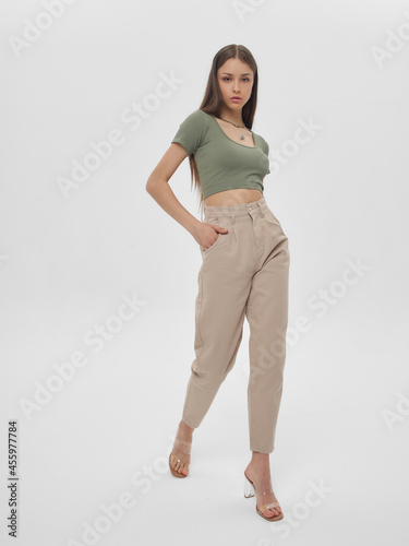 Young elegant woman with long straight hair and natural make-up in green top and beige jeans standing and posing on bright grey background