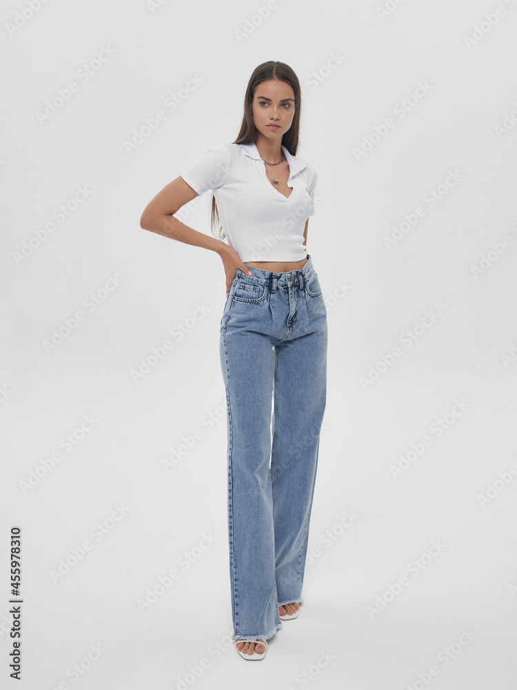 Young elegant woman with long straight hair and natural make-up in white blouse and blue jeans standing and posing on bright grey background Young elegant woman with long straight hair and natural make-up in white blouse and blue jeans standing and posing on bright grey background