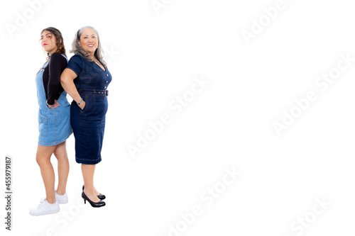 Two generations of Mexican women standing with back to back with white studio background, wide smile, light makeup, looking at camera, casual style, denim clothes. Space for text