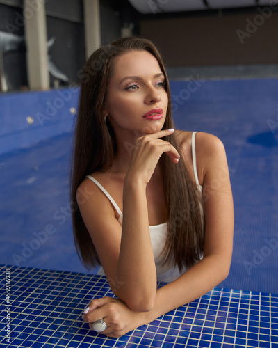 Young beautiful elegant woman wearing white mini dress and high heels standing and posing in empty pool bown with out water against blue mosaic tiles