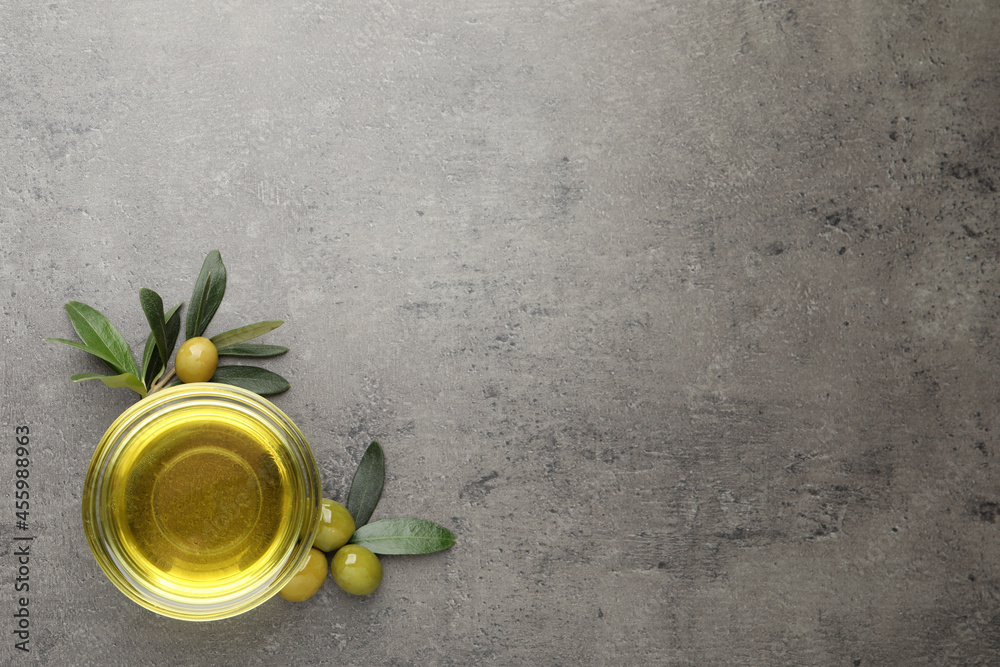 Glass bowl of oil, ripe olives and green leaves on grey table, flat lay. Space for text