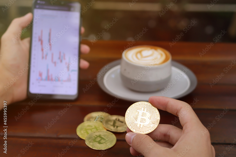 Fototapeta premium Close up of male hand holding bitcoin and mobile phone with candlestick chart on screen. Outdoor shoot in a cafe with wooden table. Cryptocurrency trading concept
