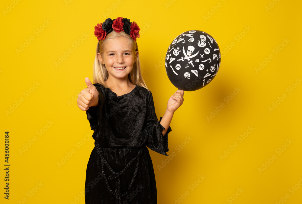 Fototapeta premium Portrait of little girl in black clothing holding black balloon with thumb up isolated over yellow background.