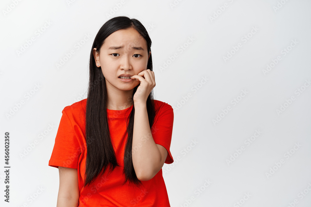 Scared asian girl biting fingers and looking at camera insecure, anxious about something, feeling worried, standing over white background Scared asian girl biting fingers and looking at camera insecure, anxious about something, feeling worried, standing over white background