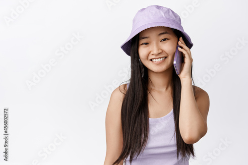 Teen asian girl talking on mobile phone, having a call and smiling at camera, happy face during conversation with smartphone, standing agaisnt white background