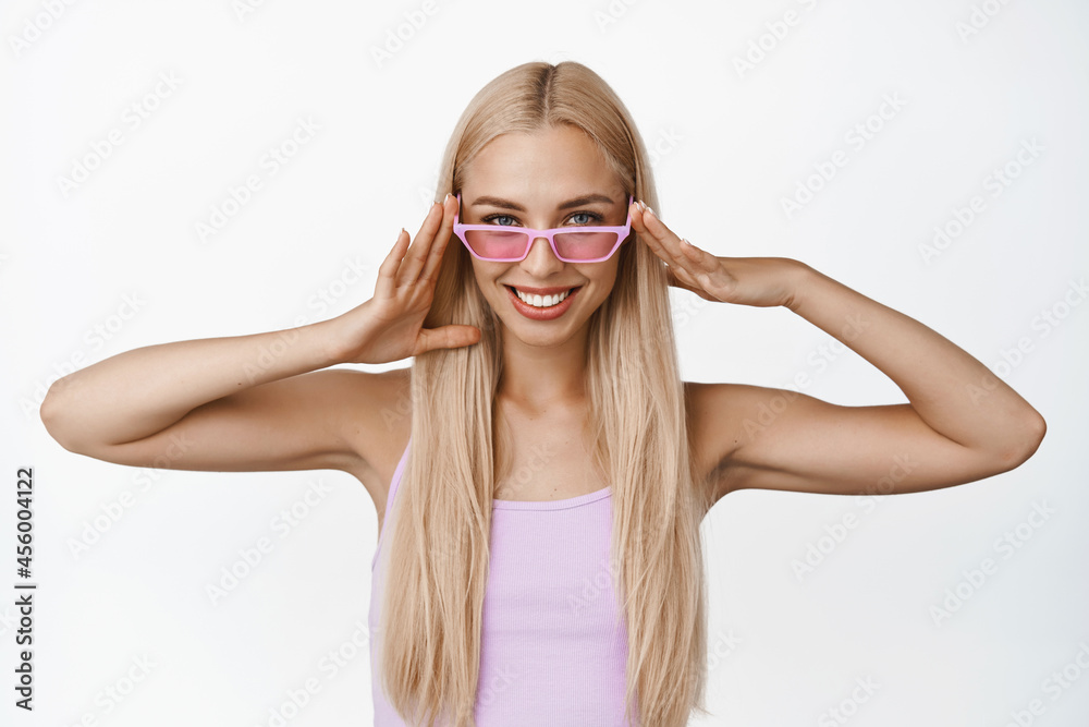 Beautiful blond girl wearing pink sunglasses and smiling at camera, concept of sunny day and vacation, standing over white background Beautiful blond girl wearing pink sunglasses and smiling at camera, concept of sunny day and vacation, standing over white background