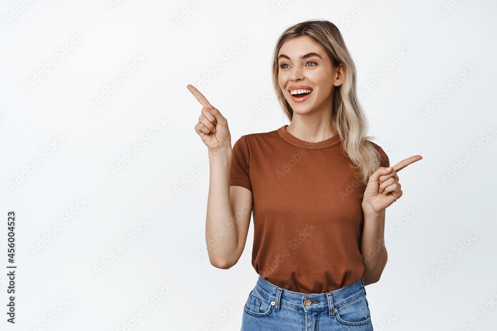 Portrait of happy young woman smiling, pointing fingers sideways and looking left at one of options, white background Portrait of happy young woman smiling, pointing fingers sideways and looking left at one of options, white background
