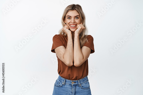 Beautiful cute woman looking at camera with fascinated face expression, holding hands on face and smiling, standing over white background