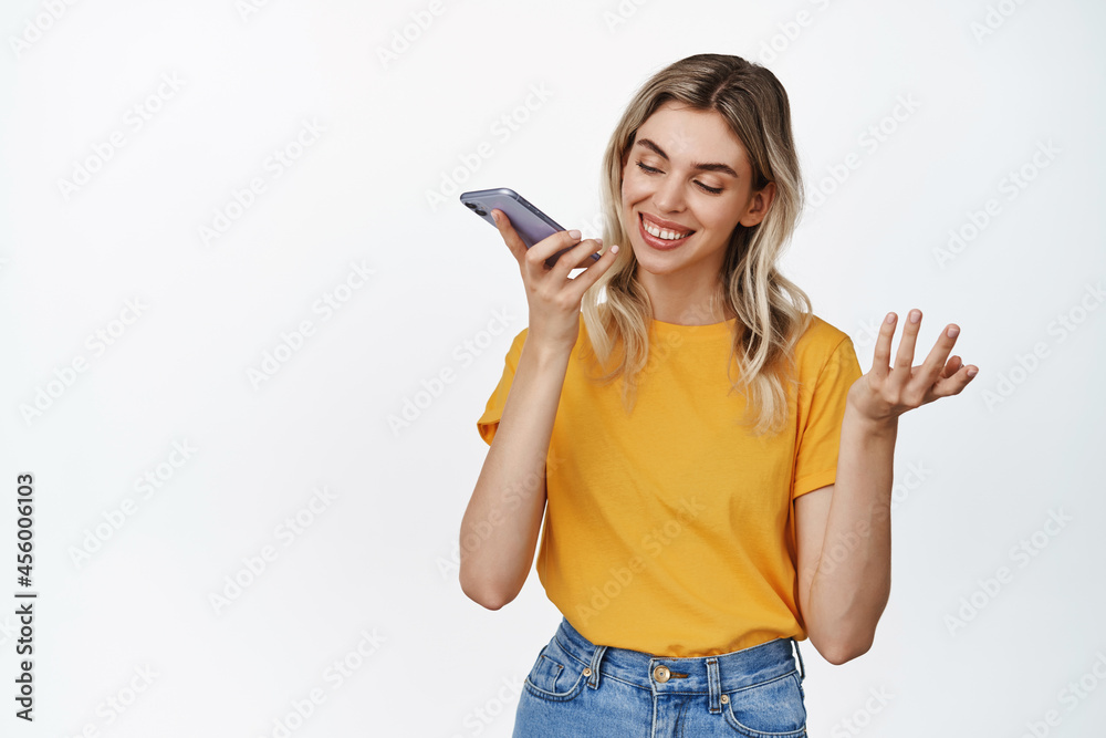 Smiling girl recording her voice, talking with speakerphone on mobile phone, record voice message or translating her words, standing over white background