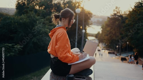 Thoughtful student doing homework in the park, thoughtfully writing notes, with laptop next to her. Dressed in a bright orange blouse, hair caught in a bun, positioned on the park strip. 4k concept