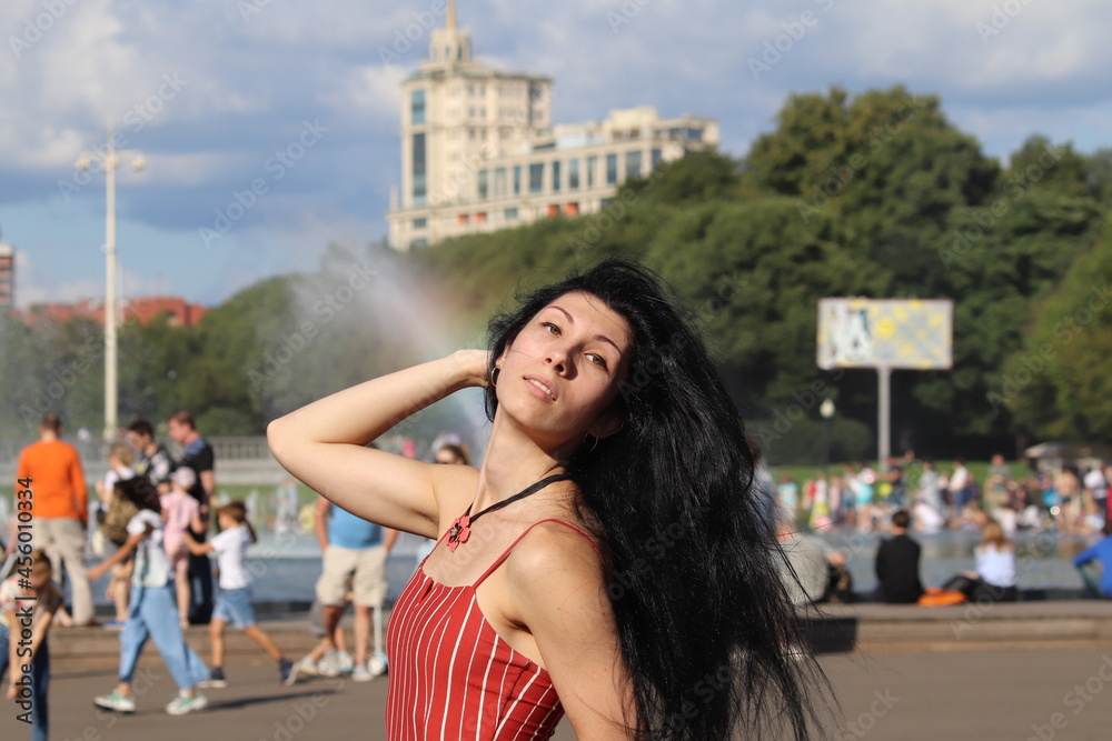 Portrait of a young woman in red dress in the city. Beautiful woman with long dark hair. Fountain and rainbow