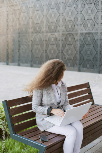 Woman working on laptop sitting on abench near modern office building photo