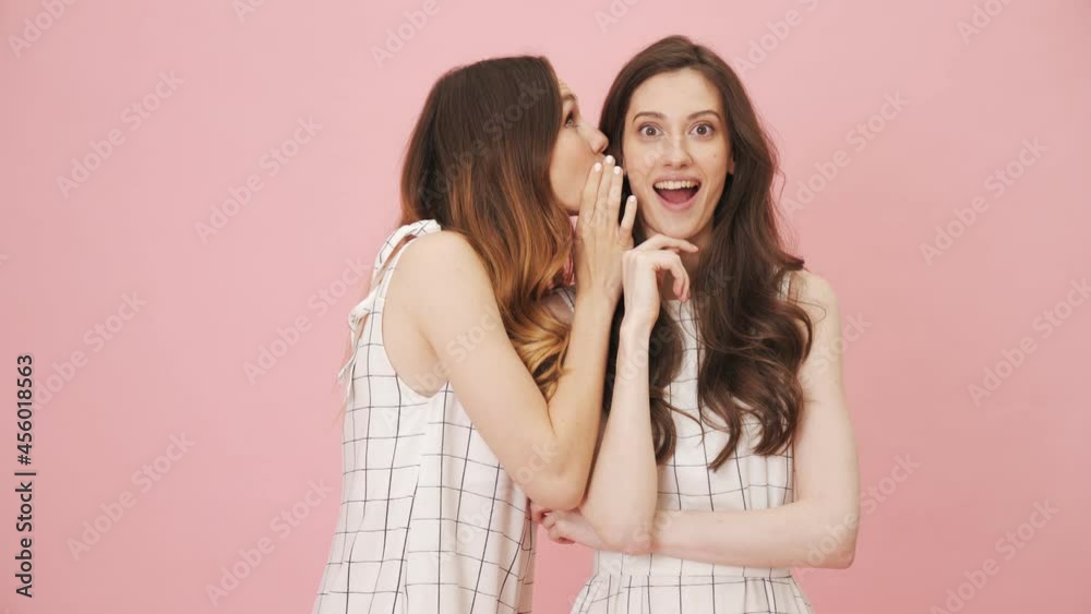 Cheerful girls sharing secrets standing isolated over pink background in the studio Cheerful girls sharing secrets standing isolated over pink background in the studio