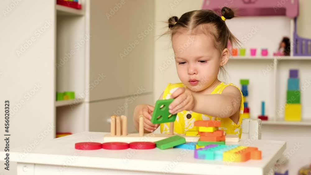 A cute little two-year-old girl is playing in the children's room with colorful toys, trying to collect the details of the game. Educational games for preschoolers. Development of hand motor skills A cute little two-year-old girl is playing in the children's room with colorful toys, trying to collect the details of the game. Educational games for preschoolers. Development of hand motor skills